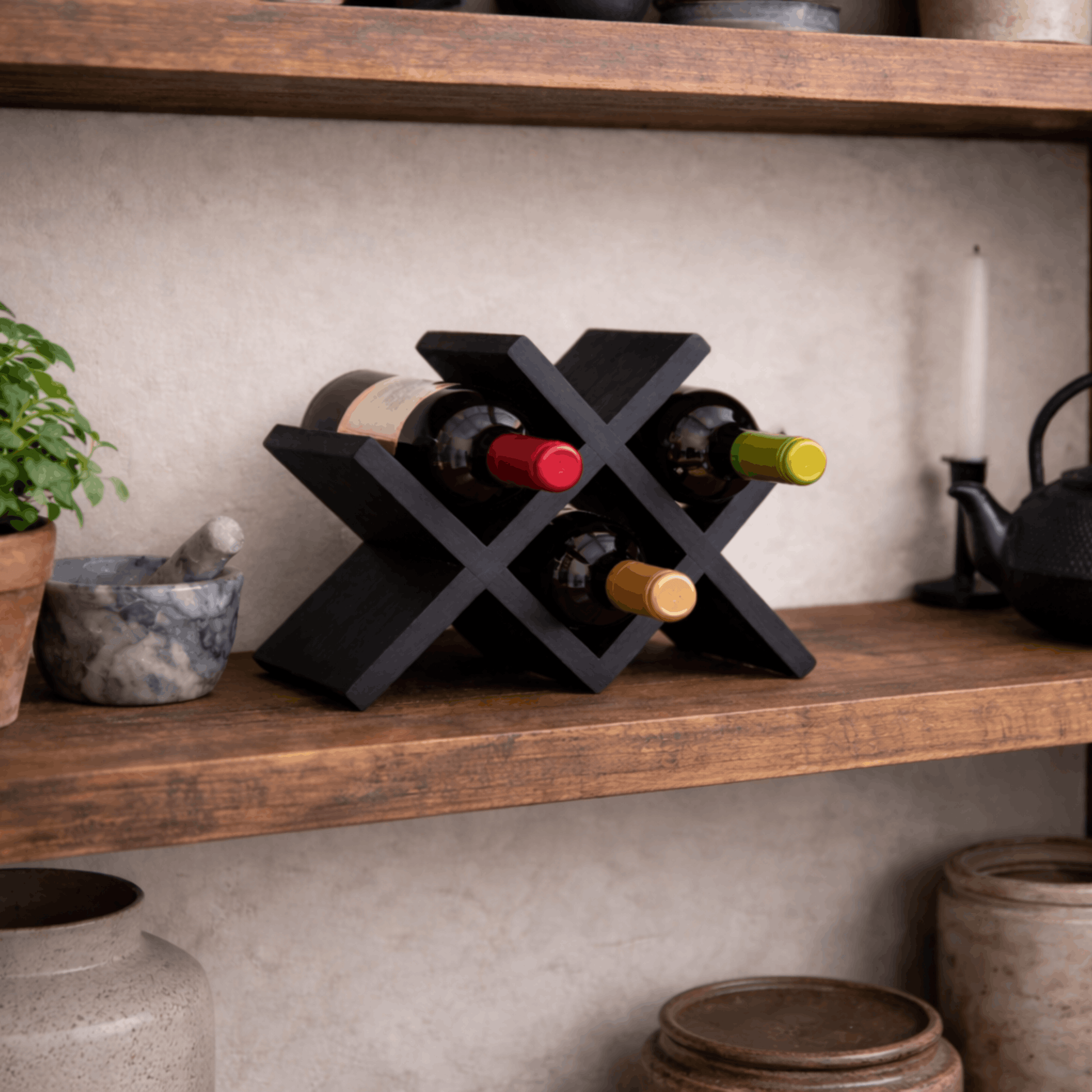 Compact zwart houten wijnrek met kruisvormig ontwerp voor vier wijnflessen, geplaatst op een houten plank in een stijlvol interieur.