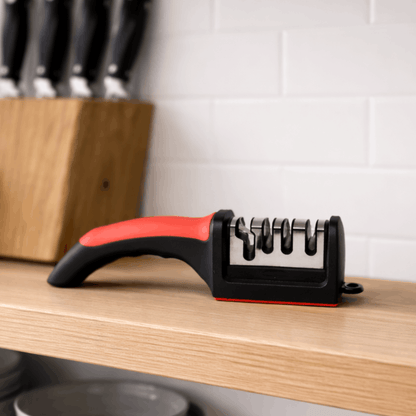 Compacte handmatige messenslijper met ergonomische handgreep en meerdere slijpsleuven, geplaatst op een keukenplank naast messenblok.