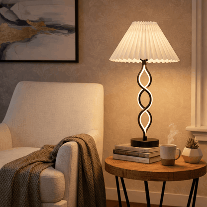 Verlichte zwarte design tafellamp met witte plissé kap op een houten bijzettafel naast een fauteuil.