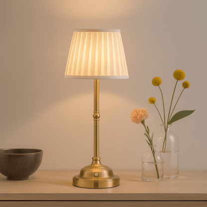 Messing tafellamp met witte plissé kap op houten tafel, verspreidt warm licht naast glazen vaasjes met bloemen.