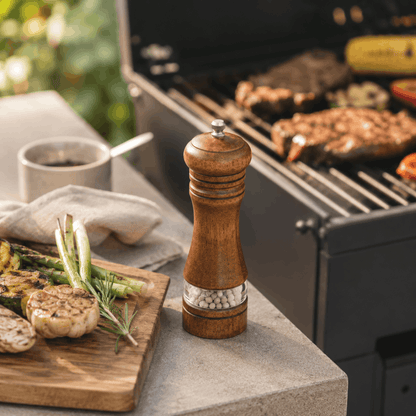 Houten peper en zoutmolen met transparant reservoir naast een barbecue met gegrilde groenten en vlees.