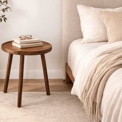 Ronde houten bijzettafel met drie poten naast een bed met boeken en een bril erop.