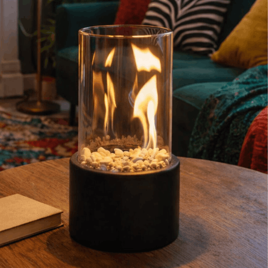 Zwarte tafelhaard met glazen cilinder en zichtbare vlammen op een houten tafel in een gezellige woonkamer.