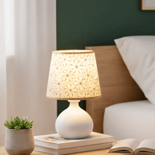 Verlichte tafellamp met bloemmotief kap op een nachtkastje naast een bed en een open boek.