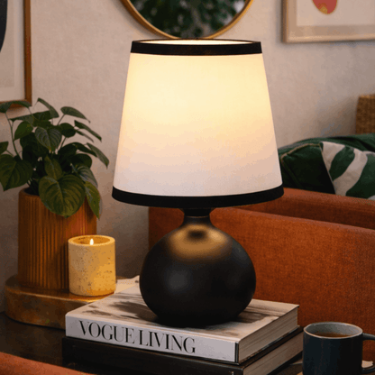 Tafellamp met ronde zwarte voet en witte kap op een stapel boeken in een gezellige woonkamer.