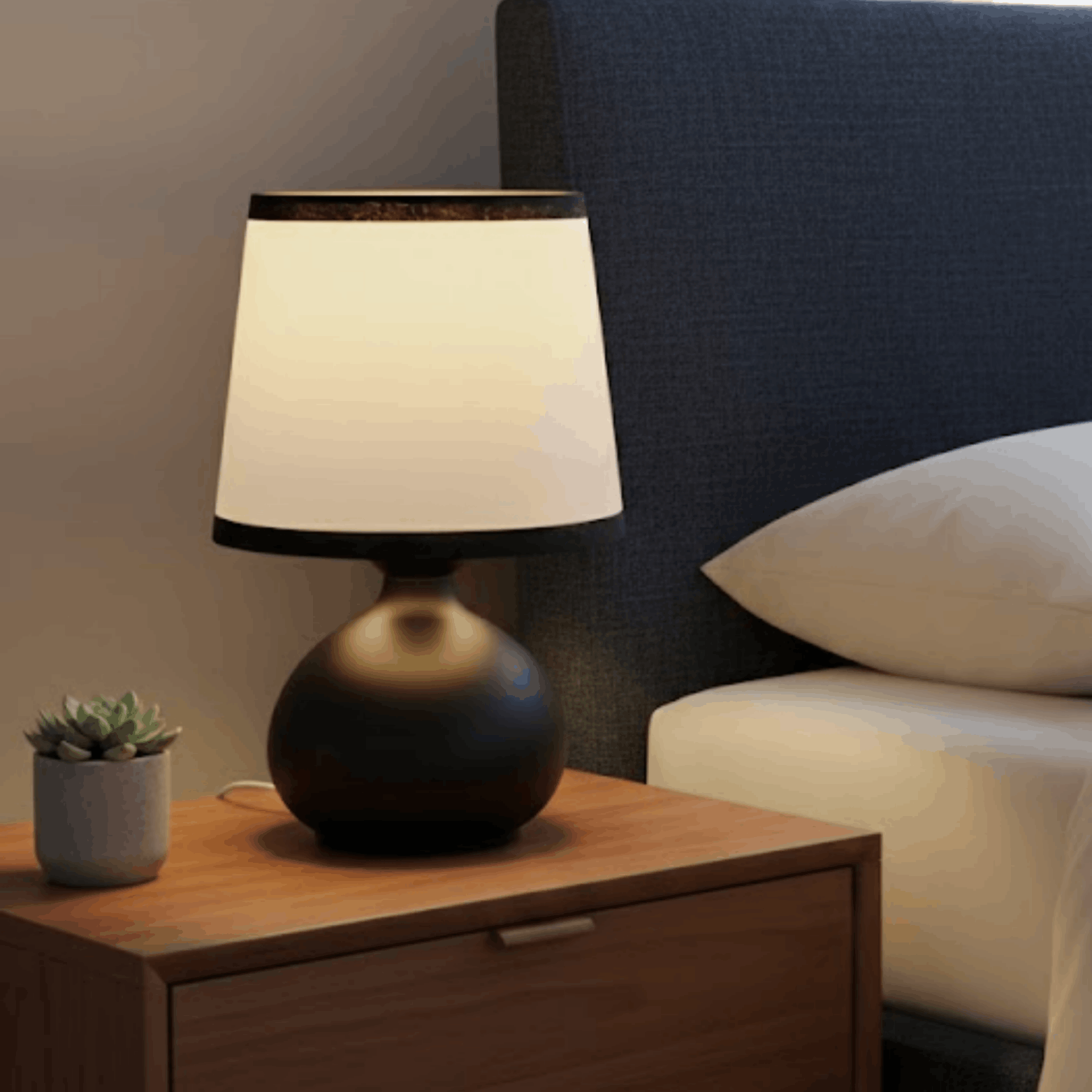 Tafellamp met zwarte bolvormige voet en witte kap op een houten nachtkastje naast een bed.