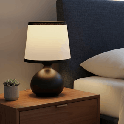 Tafellamp met zwarte bolvormige voet en witte kap op een houten nachtkastje naast een bed.