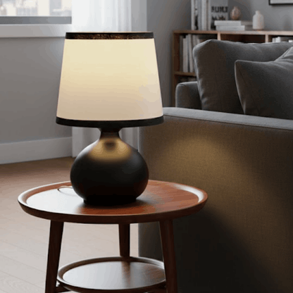 Tafellamp met zwarte ronde voet en witte kap op een houten bijzettafel naast een bank.