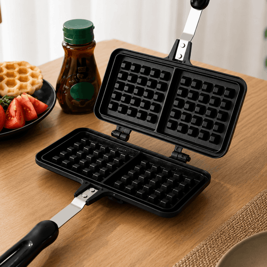 Zwart wafelijzer met dubbele wafelplaat opengeklapt op houten tafel naast siroop en wafels.