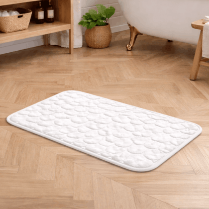 Zachte witte badmat met steenstructuur en antislip onderkant, geplaatst op een houten vloer naast een bad.
