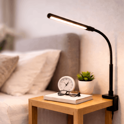 Zwarte bureaulamp met flexibele arm en smalle lichtbalk, bevestigd aan een nachtkastje naast een bed voor gericht licht.