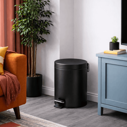 Zwarte cilindrische prullenbak met pedaal en deksel geplaatst in een woonkamer naast een kast en bank.