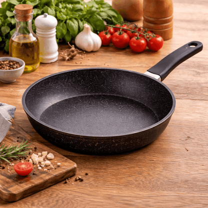 Zwarte koekenpan met antiaanbaklaag en lange handgreep, geplaatst op een houten aanrecht met ingrediënten eromheen.