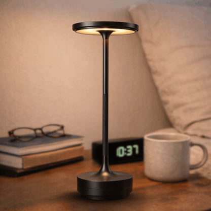 Moderne zwarte tafellamp met slanke voet en ronde lichtbron, geplaatst op een nachtkastje naast een mok en boeken.